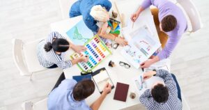 A top-down view of five diverse individuals seated around a round table, collaborating on various documents and materials. One person holds a colour chart, another reviews a graph-filled report, while others write in notebooks or point at papers. The table holds items such as pens, glasses, a planner, and a coffee mug, with a light wooden floor in the background.
