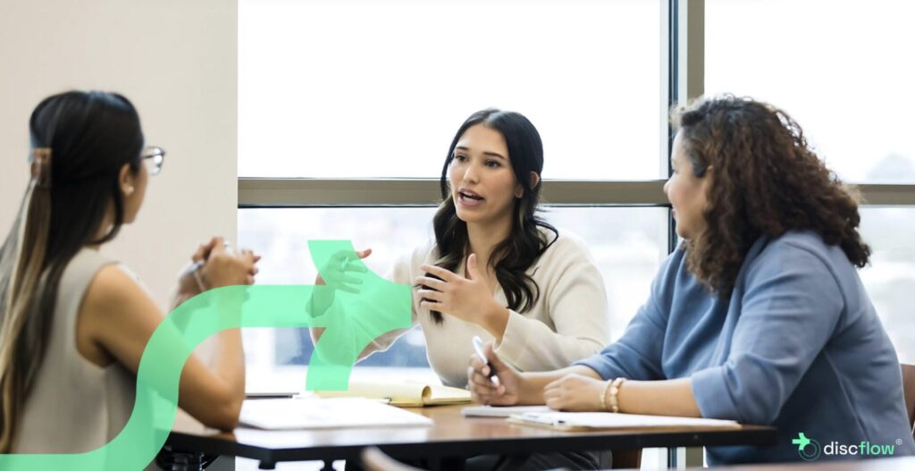 Three professional women engaged in a collaborative discussion at a table in a bright office setting, with notebooks and pens in front of them. One woman speaks while the others listen attentively. A translucent green arrow graphic and the Discflow logo are overlaid on the image.