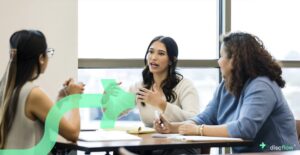 Three professional women engaged in a collaborative discussion at a table in a bright office setting, with notebooks and pens in front of them. One woman speaks while the others listen attentively. A translucent green arrow graphic and the Discflow logo are overlaid on the image.