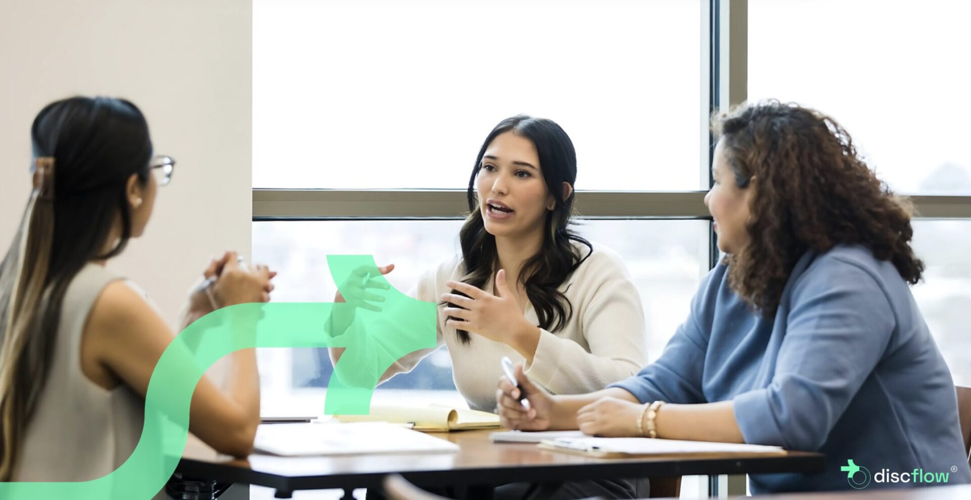 Three professional women engaged in a collaborative discussion at a table in a bright office setting, with notebooks and pens in front of them. One woman speaks while the others listen attentively. A translucent green arrow graphic and the Discflow logo are overlaid on the image.